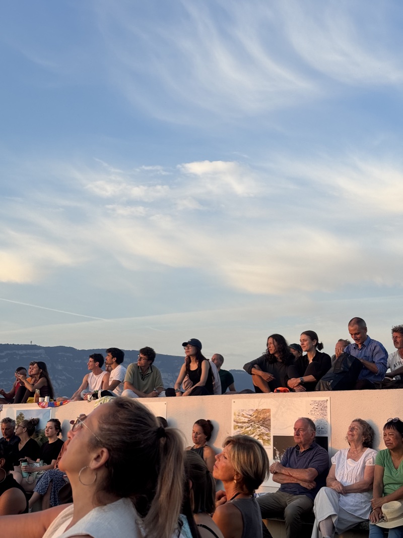 A crowd seated on a low wall at dusk, looking up at the sky, with hills behind.